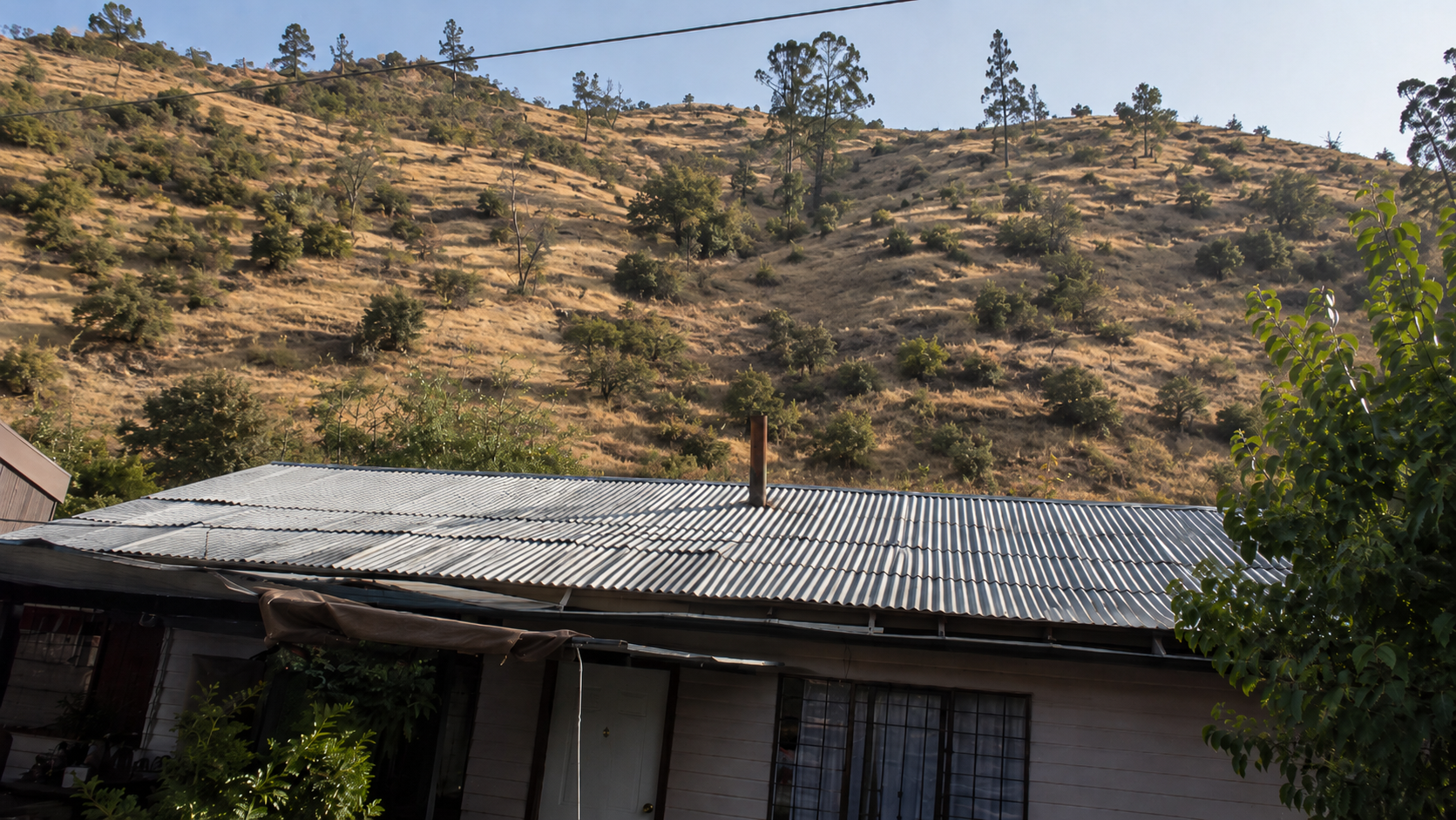 Techo de vivienda popular chilena con vegetación del cerro visible al fondo en zona de interfaz urbano-forestal